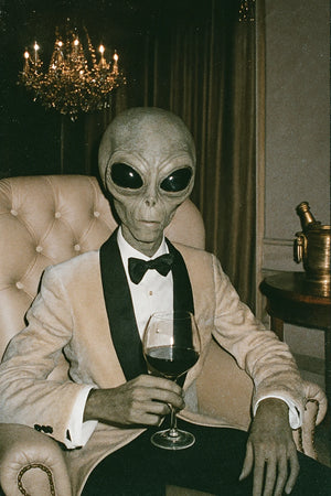 Person in an alien costume sitting in a room with a chandelier and table.