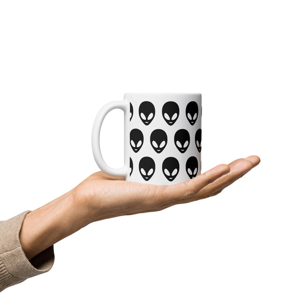 White mug with black alien face pattern held in a hand on a white background